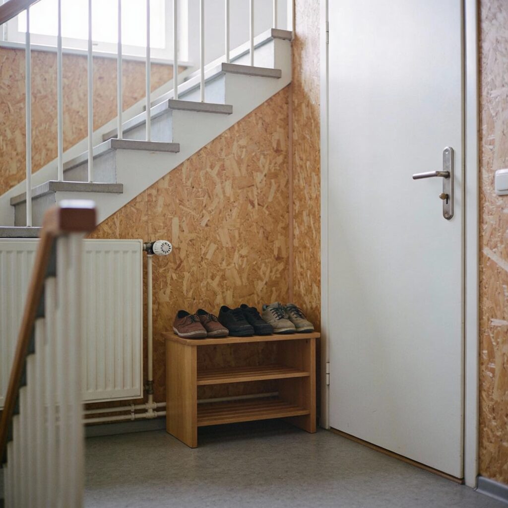 Ein kleines Holzregal mit Alltagsschuhen steht in einem engen Hausflur neben einer geschlossenen Wohnungstür.